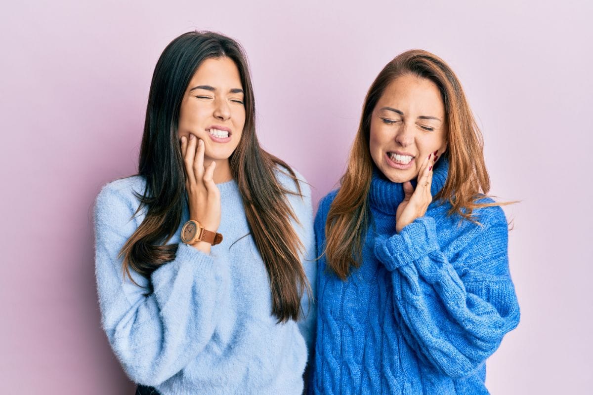 If Your Tooth Hurts on a Holiday Two smiling women in blue sweaters touching their faces against pink background.