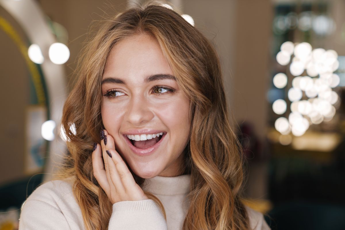 5 Ways to Smile More This Holiday Season Young woman smiling at camera, hand to face, warm indoor setting with bokeh lights background.